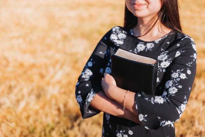 Young Smiling Christian Teenage Woman Holding A Bible Under Her Arm In The Field. Sola Scriptura. Copy Space