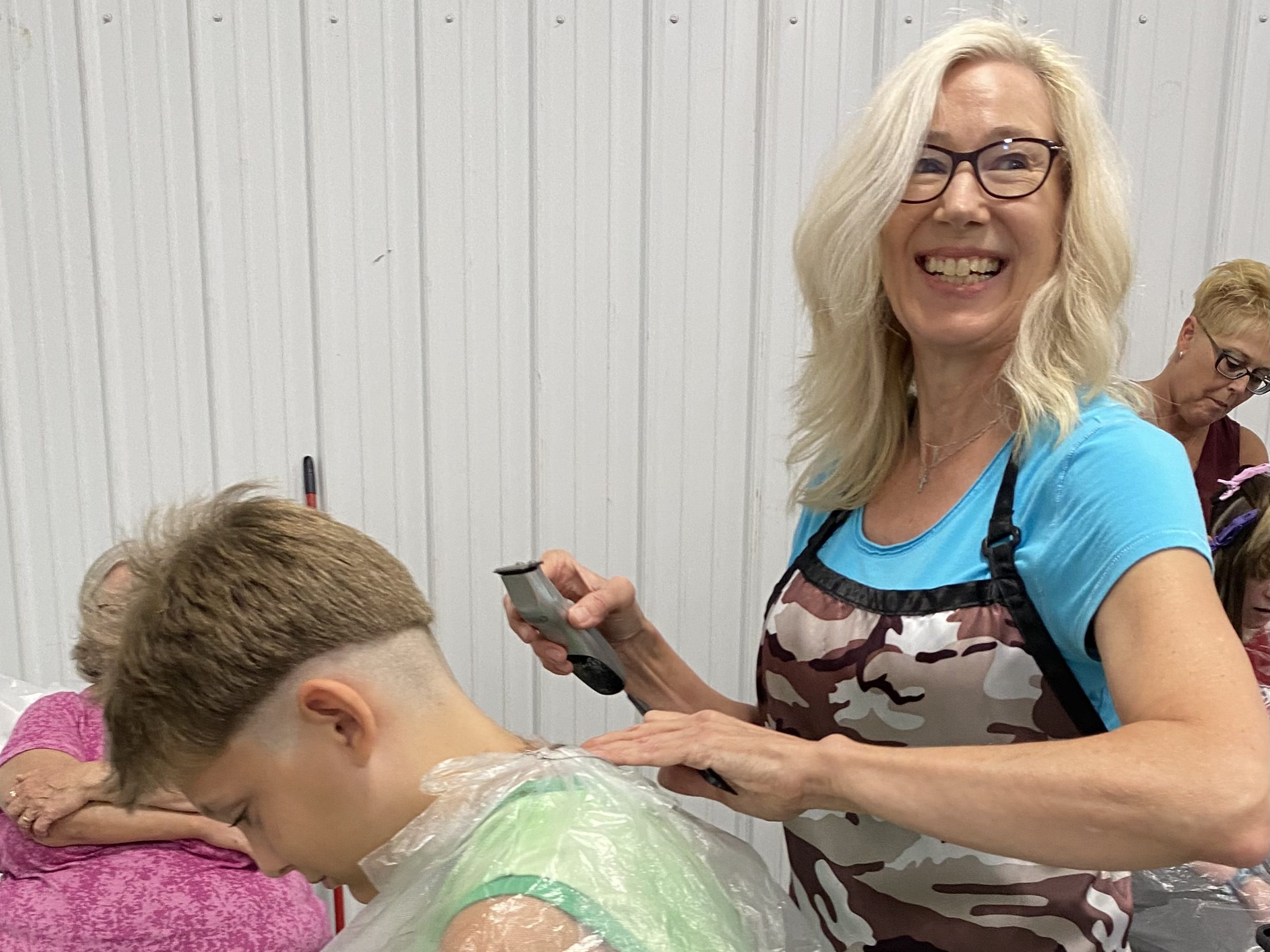 A woman wearing an apron smiles as she uses an electric razor to cut a young boy's hair.