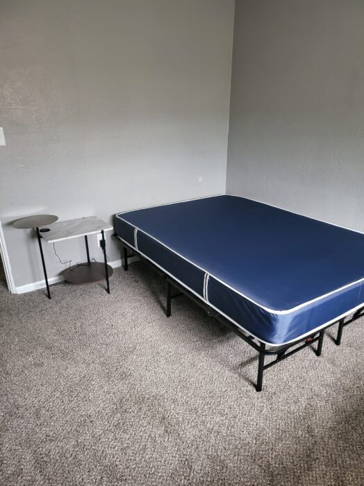 Photo shows a new blue mattress next to a small end table.