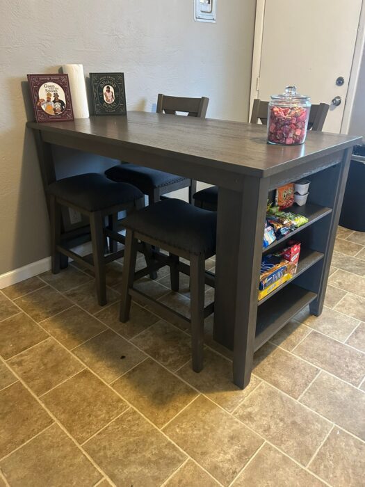Photo shows a new kitchen table with two chairs and two stools. The end of the table has shelves fully stocked with snacks.