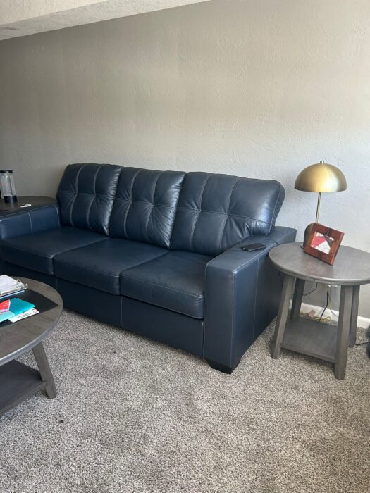 Photo shows a dark leather couch next to a small end table holding a lamp and picture frame.